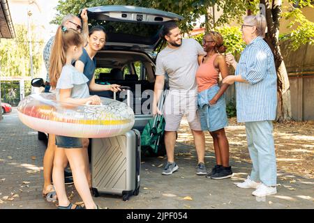 Verschiedene Familien und Freunde reisen im Urlaub, bereiten sich auf die Abreise in den Sommerurlaub und laden Gepäck in den Kofferraum. Menschen sitzen in der Auffahrt, um zum Abenteuer am Meer zu gehen. Stockfoto