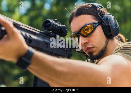 Weißer Mann mit Bart in Schutzbrille und Kopfhörer mit Zielen Maschinengewehr. Reichweite im Freien. Horizontale Aufnahme. Hochwertige Fotos Stockfoto