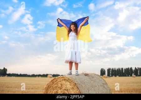 Betet für die Ukraine. Mädchen mit ukrainischer Flagge steht auf einer Heubank. Kind ohne Krieg. Schließt den Himmel über der Ukraine. Tag Des Sieges. Stockfoto