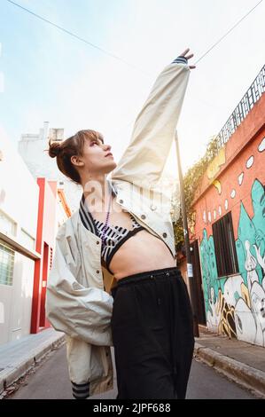 Waist up vertikales Porträt einer trans jungen lateinischen kaukasischen Frau, die auf der Straße zum Himmel zeigt und nach oben schaut. Stockfoto