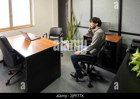 Ein Geschäftsmann, der in seinem Büro sitzt und am Telefon telefoniert, schaut pensiver aus dem Fenster Stockfoto