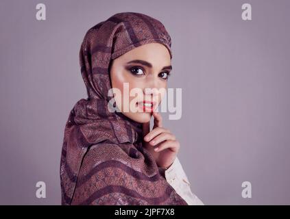 Lippen so rot wie ein guter Wein. Studioporträt einer selbstbewussten jungen Frau, die einen farbenfrohen Kopftuch trägt, während sie einen roten Lippenstift auf ihre Lippen aufträgt. Stockfoto