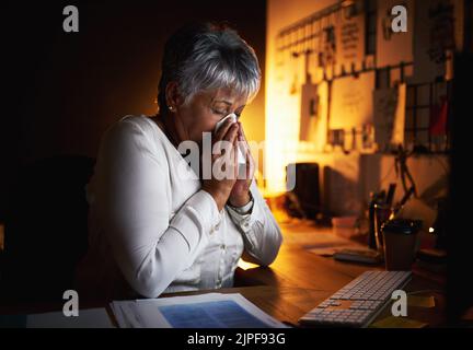 Ich hoffe wirklich, dass ich diesmal nicht die Grippe erwischt habe. Eine reife Geschäftsfrau bläst sich die Nase, während sie spät in einem Büro arbeitet. Stockfoto