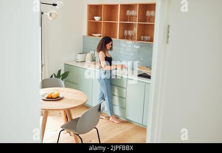 Junge Frau, die in der Küche Tee, Kaffee oder Morgengetränk zubereitet, während sie sich zu Hause entspannt. Frau genießt, bereitet und trinkt ein Getränk für eine gemütliche Stockfoto
