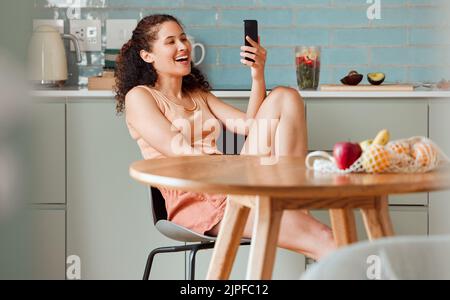 Frau, die am Telefon unterwegs ist und mit einer Online-App einen Beitrag in den sozialen Medien liest, während sie sich in der Küche entspannt. Frau entspannt, lächelt und genießt einen lustigen Beitrag Stockfoto