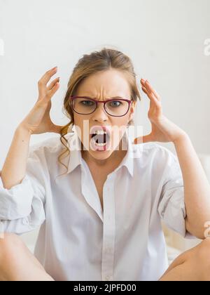 Porträt einer jungen wütenden Frau, die die Hände in wütender Geste hält. Junge Hündin mit roten Haaren in weißem Hemd. Negative menschliche Emotionen, Gesichtsausdrücke. Stockfoto