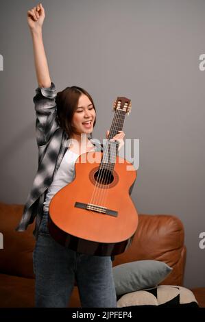 Glückliche und talentierte junge asiatische Musikerin, die ihre Akustikgitarre hält und ihr Glück durch Heben ihrer Hand zum Ausdruck bringt. Stockfoto