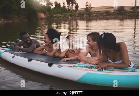 Freunde, Urlaub und Spaß haben, während Sie sich auf ein Paddle-Board lehnen und in einem See reden. Glückliche und vielfältige Menschen lachen, während sie das Wasser genießen und Stockfoto