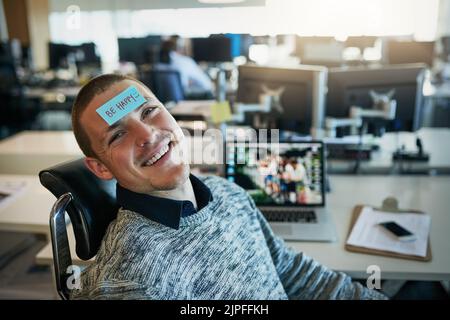 Er ist immer so glücklich, wie er sein kann. Ein fröhlicher junger Geschäftsmann mit einer Haftnotiz auf der Stirn, der sagt, sei glücklich, während er im Büro sitzt. Stockfoto