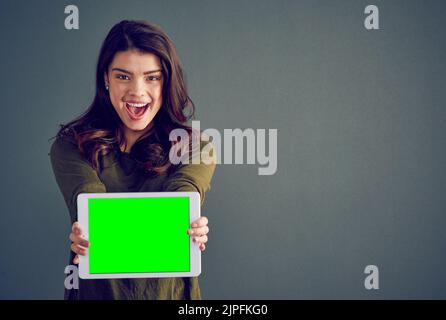 Endlich habe ich auch meine bekommen. Studioaufnahme einer fröhlichen jungen Frau, die ein digitales Tablet hält und vor einem dunklen Hintergrund steht. Stockfoto