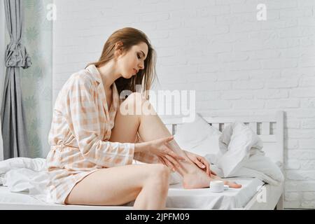 Junge Frau im Pyjama auf dem Bett sitzend und Körpercreme auftragen. Frau trägt nach dem Epilieren oder Enthaarung eine Bademantel-Reinigungslotion. Stockfoto