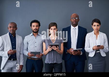 Vermarkten Sie Ihr Unternehmen auf technologische Weise. Studio-Aufnahme einer Gruppe von Geschäftsleuten, die verschiedene Geräte vor grauem Hintergrund verwenden. Stockfoto