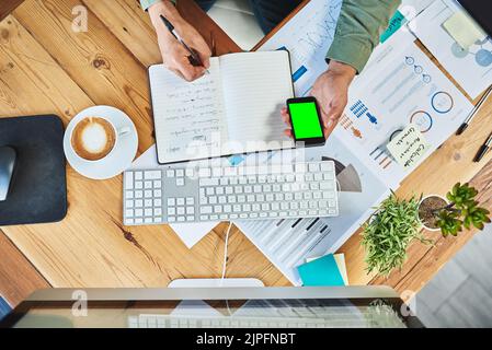 Schreiben Sie alle wichtigen Dinge auf. High-Angle-Aufnahme eines unerkennbaren Geschäftsmannes, der ein Notebook und ein Mobiltelefon in der Hand hält, während er hinter seinem Schreibtisch arbeitet Stockfoto