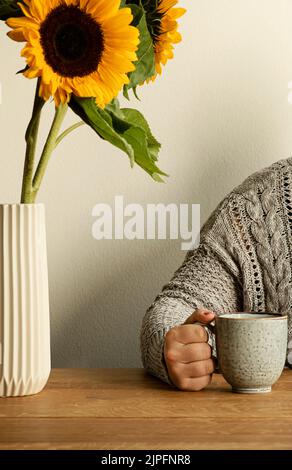 Stillleben mit schönen Sonnenblumen in einer Vase und unkenntlich Frau in einem gemütlichen Strickpullover Kaffee trinken an Holztisch. Stockfoto