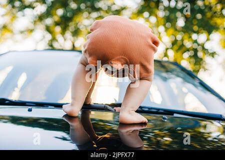 Baby versucht, auf der Motorhaube des Autos auf der Seite der Straße geparkt stehen. Sommerurlaub Spaß. Schönes junges Mädchen. Frohe Familie. Stockfoto