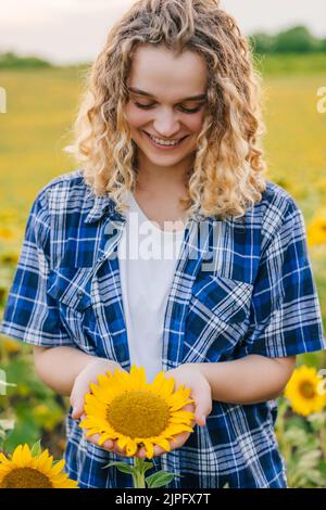 Lockige Bäuerin, die mitten auf einem Sonnenblumenfeld steht, eine Sonnenblume in der Hand hält und sie mit Bewunderung anschaut. Landarbeiter Stockfoto