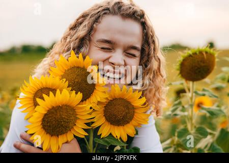 Glücklich lockiges Mädchen umarmt Sonnenblumenblumen in einem Feld von Sonnenblumen bei Sonnenuntergang. Schönheitsporträt. Natürliche Sommerlandschaft. Agronom und Landwirt Stockfoto