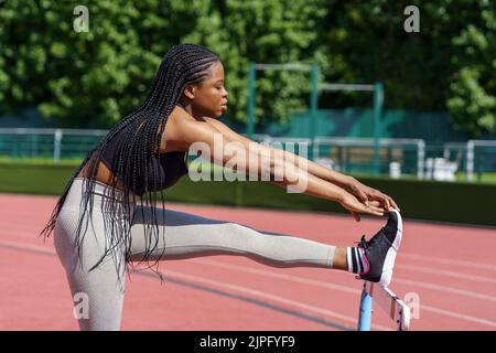 Schwarze junge Frau mit langen Zöpfen streckt Beine und Körper mit Hürden Barriere auf Sportplatz Stockfoto