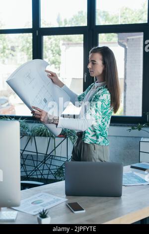 Seitenansicht eines queeren Designers, der die Blaupause in der Nähe von Geräten im Büro hält Stockfoto