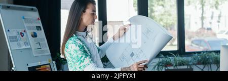 Seitenansicht der nichtbinären Person, die Blaupause in kreativer Agentur hält, Banner Stockfoto