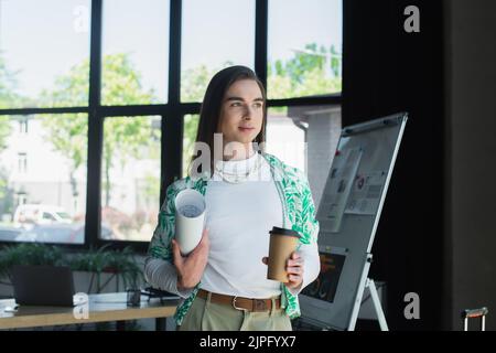 Brünette queer Person hält Blaupause und Kaffee im Büro zu gehen Stockfoto