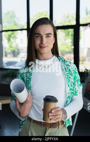 Lächelnder, queerer Designer, der Blaupause und Kaffee hält, um ins Büro zu gehen Stockfoto