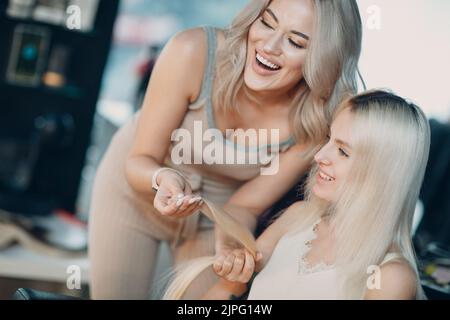 Friseur Frau macht Haarerweiterungen für junge Frau mit blonden Haaren im Schönheitssalon. Professionelle Haarverlängerung Konzept Stockfoto