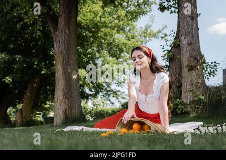 Stilvolle Frau, die im Park Orangen in einem Strohsack auf einer Decke ansieht Stockfoto