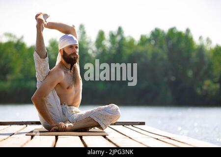 Junger Mann praktiziert Yoga am Flussufer bei Sonnenaufgang, im Freien. Selbstbewusstsein und Akzeptanz. Konzept der psychischen Gesundheit, Entspannung, Harmonie mit Stockfoto