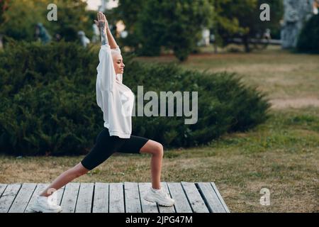 Porträt einer jungen, lächelnden, tausendjährigen europäischen Kurzhaarigen, die im Park Yoga macht. Schöne glücklich blonde Mädchen im Freien. Stockfoto