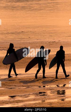 Eine Gruppe von Surfern, die ihre Surfbretter tragen und am Fistral Beach entlang spazieren, der von einem intensiven Sonnenuntergang in Newquay in Cornwall in Großbritannien umrast wird. Stockfoto