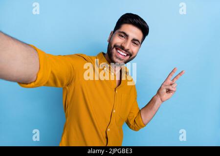 Foto von schönen toothy strahlenden Kerl gekleidet gelben Hemd zeigt V-Zeichen lächelnd tun Selfie isoliert auf blauem Hintergrund Stockfoto