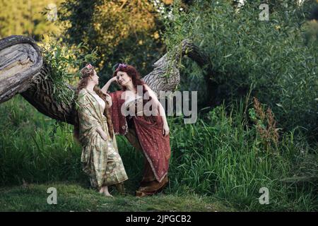 Porträt von zwei schönen reifen Frauen in einem Nymphen-Stil Kleid gekleidet. Frauen sprechen in der Nähe des Stammes eines gefallenen Baumes. Stockfoto
