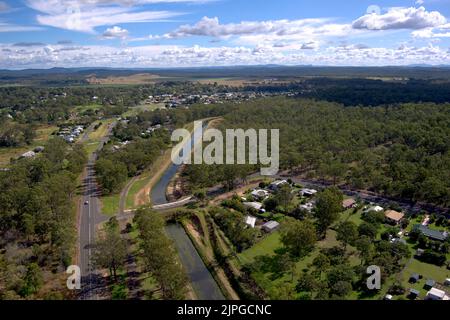 Aus der Vogelperspektive auf eine gekrümmte Straße und einen Bewässerungskanal durch eine Vorstadtlandschaft mit Häusern und Grün unter einem blauen Himmel mit Wolken Stockfoto