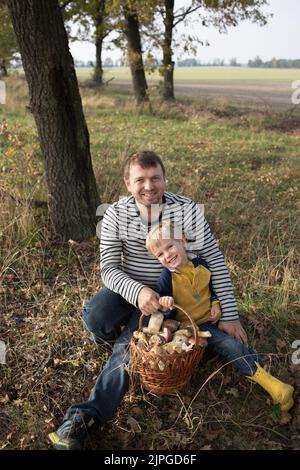 Fröhlicher Vater und Sohn sitzen auf einem Baumstumpf im Wald und halten einen Korb aus Weidenstroh, gefüllt mit essbaren Pilzen. Hobby zum Sammeln von Pilzen. selecti Stockfoto