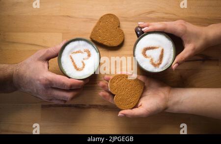 Männliche und weibliche Hände halten identische Tassen Kaffee mit Milch, in denen Zimt-Herzen sichtbar sind. In der Nähe befinden sich zwei herzförmige Lebkuchenkekse. Stockfoto