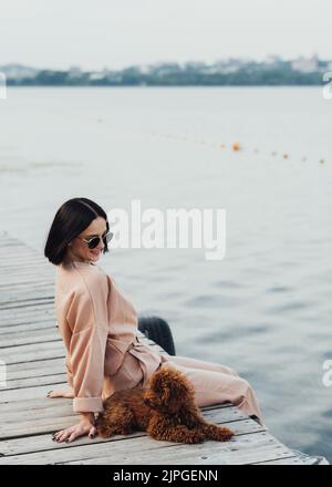 Brünette Frau sitzt auf Pier mit ihrem rothaarige Hund Spielzeug Pudel Stockfoto
