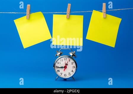 Eine Uhr neben einem Seil mit gelben Haftnotizen, die unten hängen, um Ziele, Erinnerungen, wichtige Termine oder Geburtstage zu enthalten. Blauer Hintergrund. Zeit zum erinnern Stockfoto