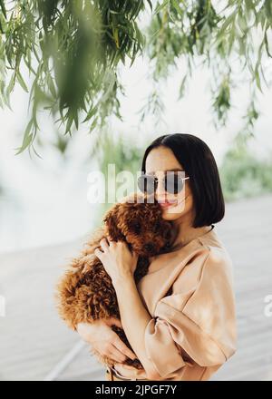 Junge Brünette Frau hält sich an den Händen und umarmt ihren kleinen rothaarigen Hund Rasse Spielzeug Pudel Stockfoto