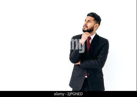 Rätselhafter, nachdenklicher, eleganter, einflussreicher arabischer oder indischer Mann im formellen Anzug, stehen auf isoliertem weißem Hintergrund, blicken pensiv weg, denken, planen, träumen. Platz zum Kopieren Ihrer Präsentation Stockfoto