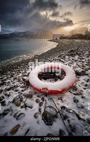 Lifebuoy auf einem Kiesstrand während eines Sonnenuntergangs im Winter auf Godøy, Sunnmøre, Møre Og Romsdal, Norwegen. Stockfoto