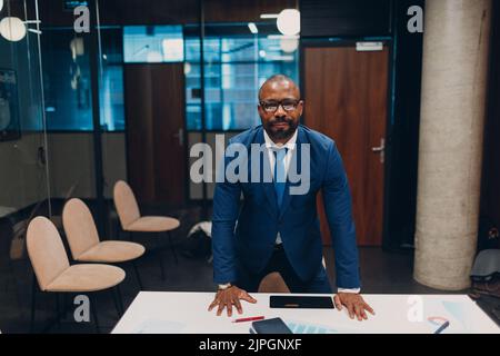 Portrait lächelnder afroamerikanischer Geschäftsmann in blauem Anzug steht am Tisch für Treffen im Büro. Stockfoto