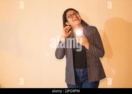 Eine kaukasische Frau in grauer Jacke, die das Telefon mit blinkenden Lichtes hält und lächelt Stockfoto