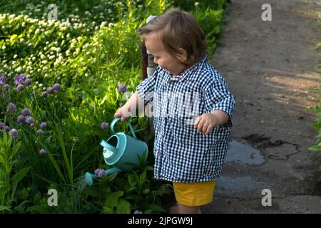 Ein kleiner Junge, der auf der Route im Garten des Hinterhofs steht, gießt Blumen aus einer Gießkannte. Das Konzept der Sommeraktivitäten im Garten, Sommersaison, Gartenarbeit, Pflanzen- und Blumenanbau. Stockfoto