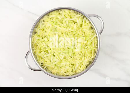 Geriebene, gesalzene Zucchini, die in einem Stahlsieb zum Ausstreten des Wassers über einem weißen Marmorbrett platziert wurden. Zubereitung von Knochenmark-Gemüse f Stockfoto