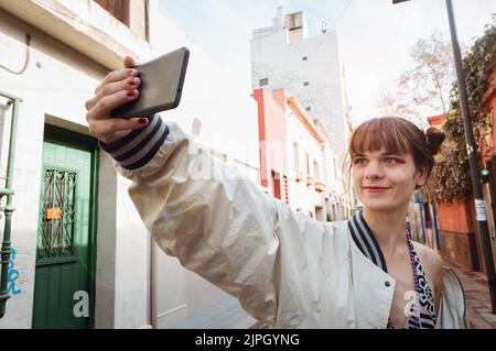 kaukasische junge lateinische Transgender-Frau mit rotem Make-up und halten Mobiltelefon mit einer Hand in städtischen Wohnstraße stehend, lächelnd unter einem Stockfoto