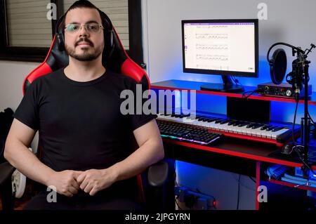 Porträt eines jungen Latino-Musikproduzenten mit kurzen Haaren, Bart und Brille, der zu Hause in seinem Musikstudio sitzt und glücklich die Kamera anschaut, wi Stockfoto