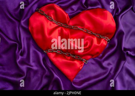 Rotes Satin Herz mit Ketten auf lila Hintergrund gebunden, Ehe Einschränkungen oder Eifersucht Metapher Stockfoto