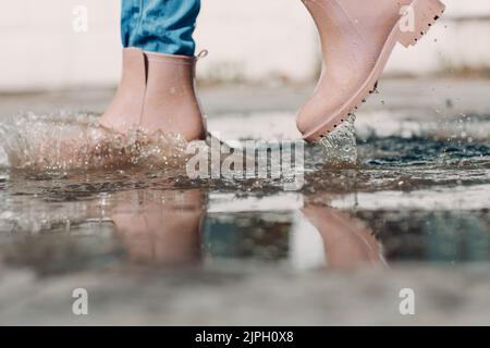 Frau mit Regengummistiefeln, die beim Herbstregen mit Wasserspritzern und Tropfen in die Pfütze laufen und springen. Stockfoto
