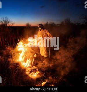 Feuerwehrmann Ökologe bei der Bekämpfung des Feuers auf dem Feld in der Nacht. Mann in Schutzanzug und Gasmaske in der Nähe brennendes Gras mit Rauch und gelbem Dreieck mit Schädel und Kreuzknochen Warnschild. Stockfoto